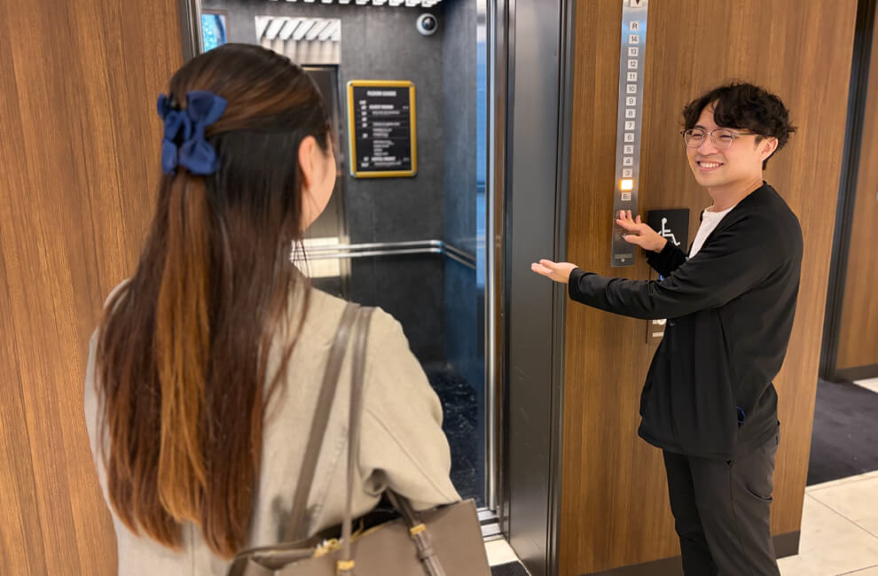 つなぐはぐくむツーリズムKidsジョブチャレンジ フロントスタッフがお客様にエレベーターをご案内しているシーン