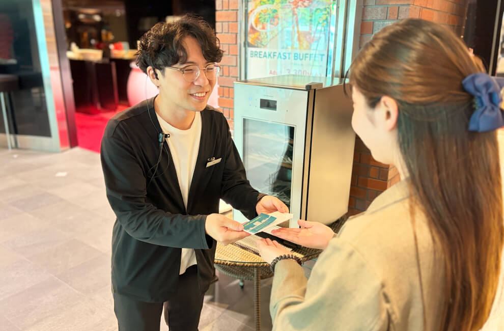つなぐはぐくむツーリズムKidsジョブチャレンジ フロントスタッフがお客様におしぼりタオルをお渡ししているシーン